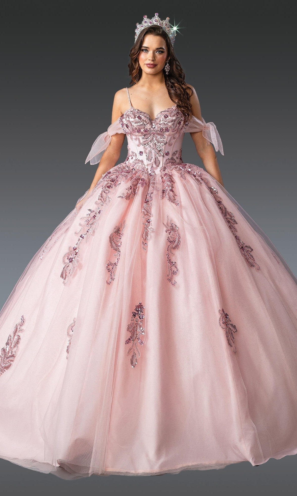 A woman wearing the Quinceanera Dress 1902 by Dancing Queen in blush, featuring a tulle ballgown skirt with sequin embellishments and an embellished bodice, paired with a matching tiara, stands against a plain gray background.